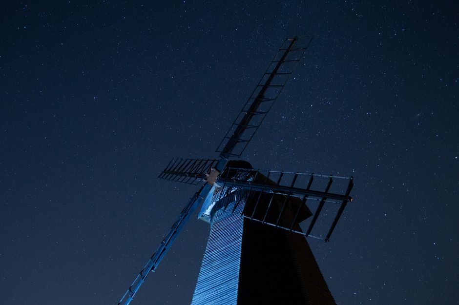 A black and white photo of a historic windmill with a cylindrical stone tower featuring a rough textured surface and small rectangular windows evenly spaced along its height. The windmill has four lar